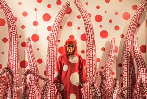 Yayoi Kusama in a polka dot dress and sunglasses, standing in front of one of her art installations