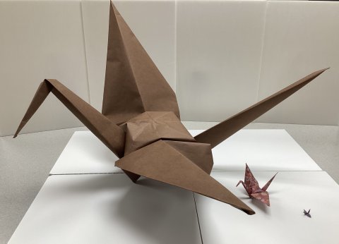A very large origami crane about 2 feet long made of brown paper, next to a regular sized origami crane made of pink floral paper, next to a tiny origami crane about an inch long made with dark purple paper, sitting on a white table with a white background.