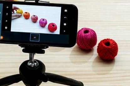 A phone attached to a mini tripod on a table pointed at small spools of red, pink, yellow thread