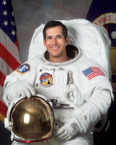 Portrait of an American astronaut, John Herrington, in a white space suit behind a US flag and a NASA flag.