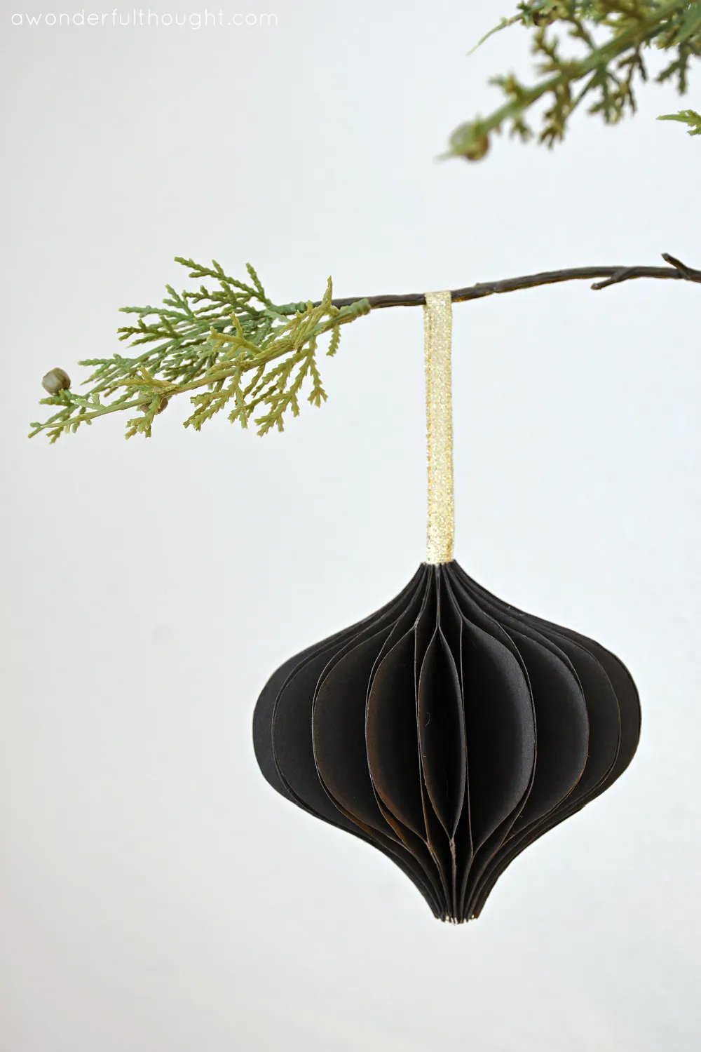 Honeycomb paper teardrop ornament with gold ribbon hanging on a tree branch. 